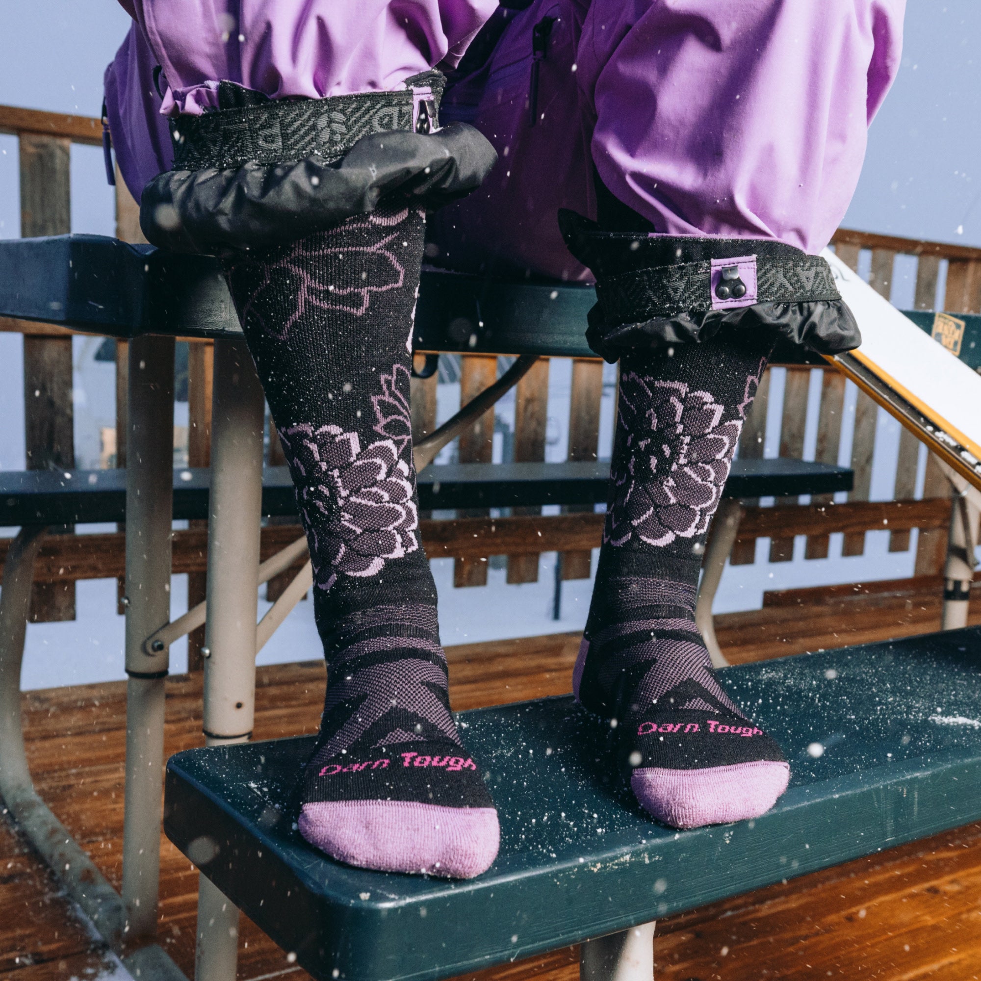 model sitting on a picnic table in the snow wearing the 8056 dahlia over the calf ski and snowboard sock in black