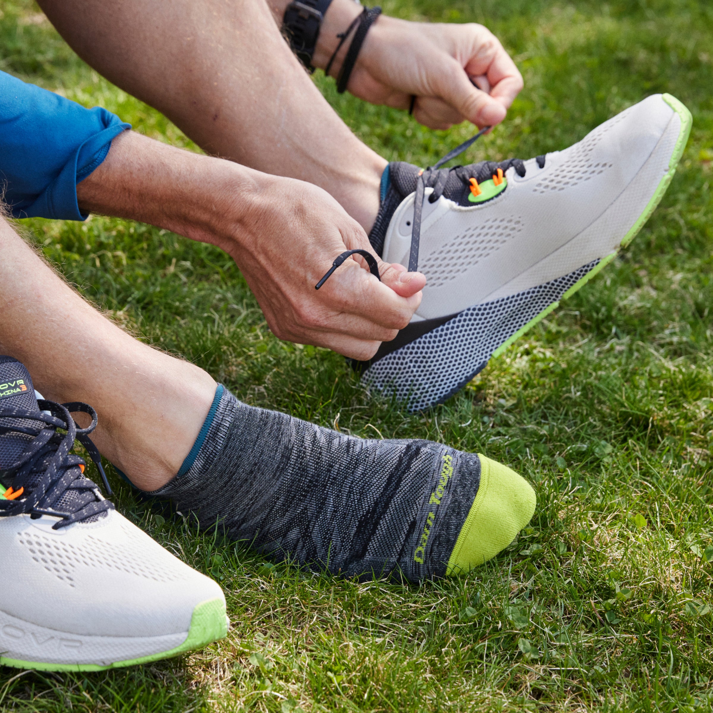 Close up shot of model sitting on grass wearing 1039 socks in comet and lacing up white running shoe on left foot.