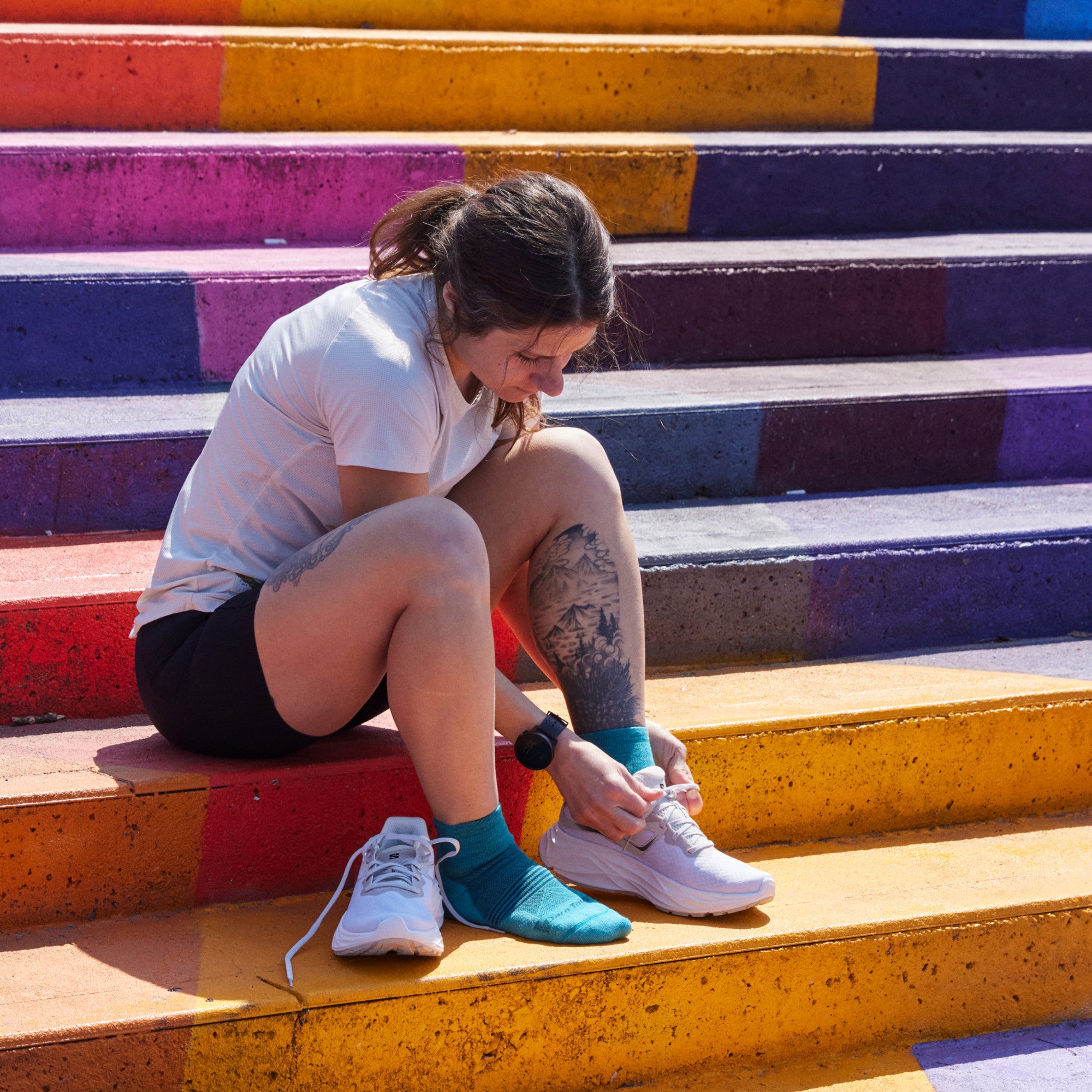 Model sitting on colorfully painted concrete steps and wearing 1113 socks in Cyan colorway and lacing up white running shoe on left foot.