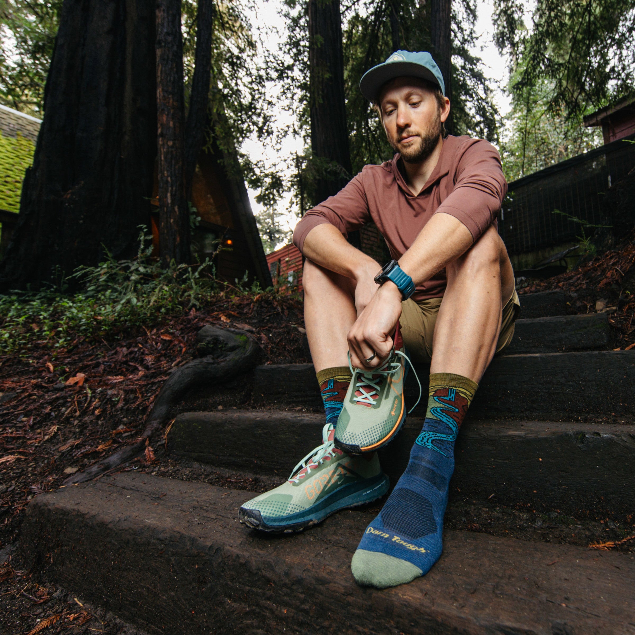 Model sitting on wooded steps putting sneaker on wearing the 5020 chasing waterfalls micro crew hiking sock in Denim