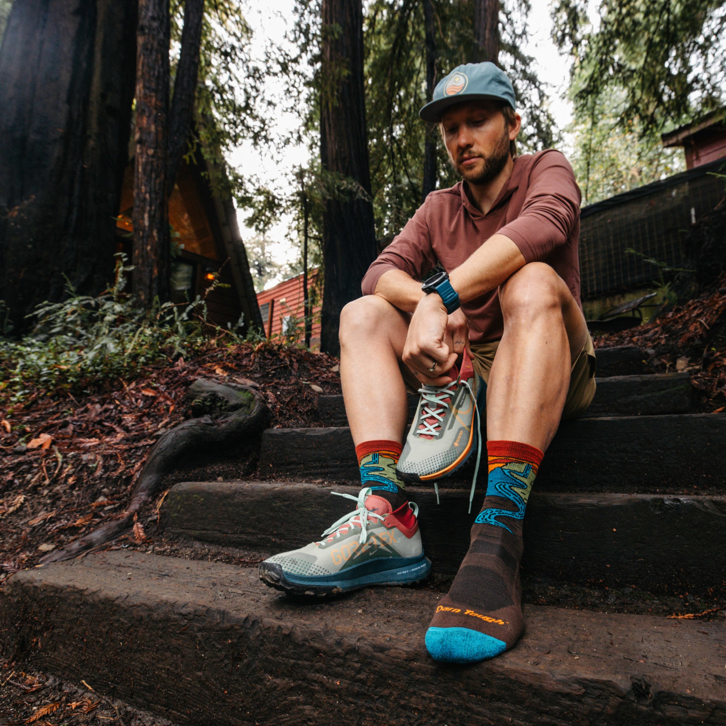 Model sitting on wooded steps putting on sneaker wearing the 5020 chasing waterfalls micro crew hiking sock in Earth