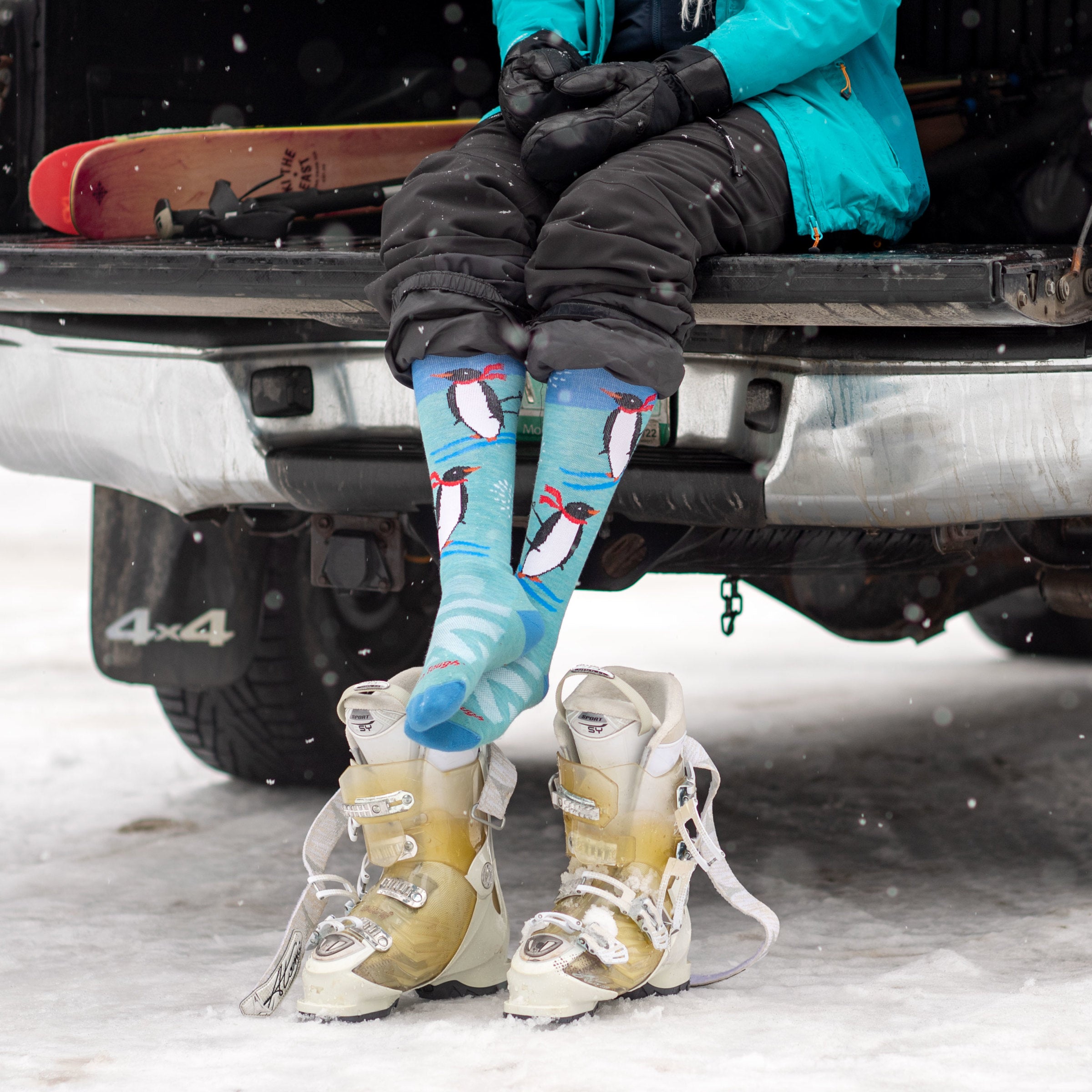 Model sitting in car trunk wearing women's penguin peak over-the-calf snow sock with white ski boots on ground