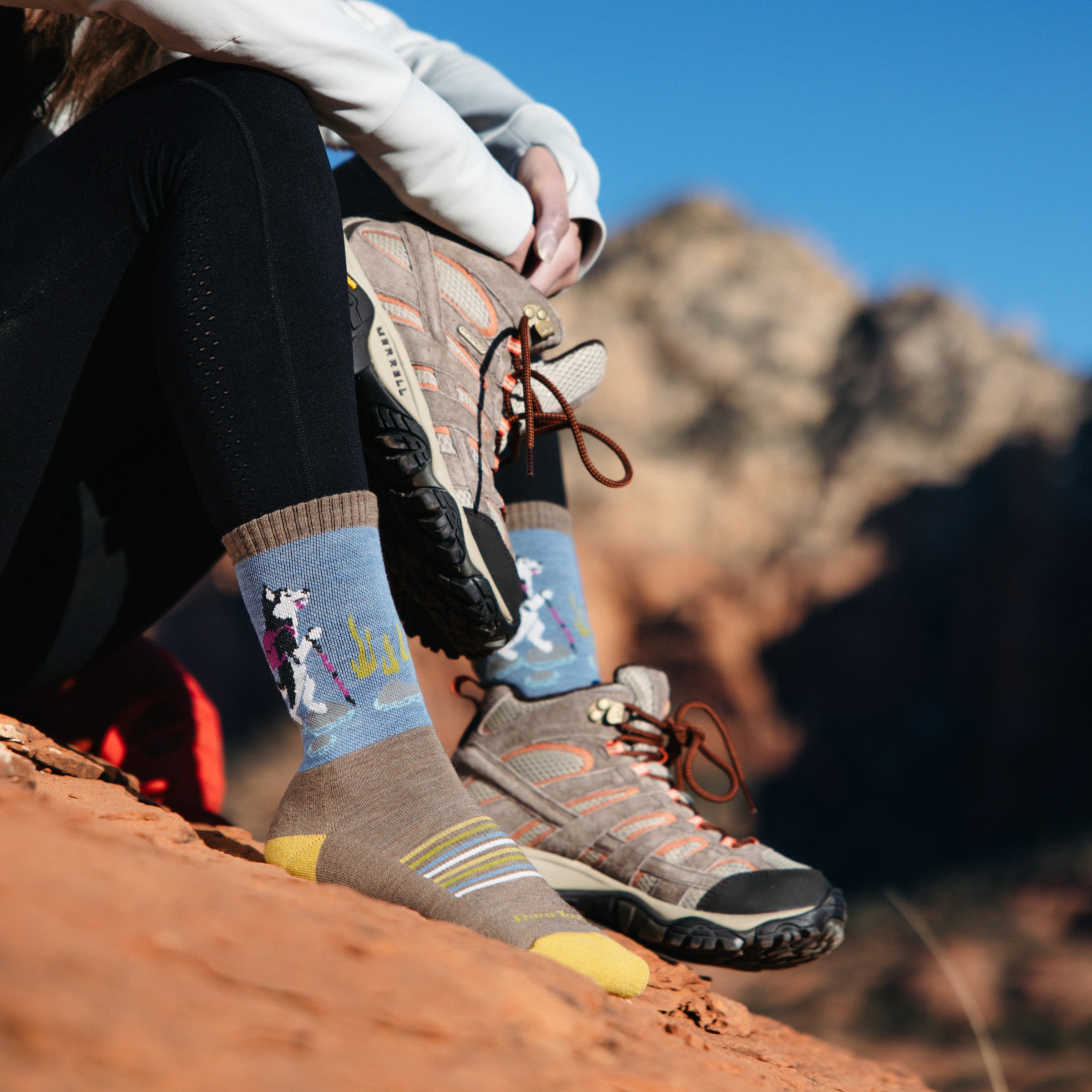 Image of model sitting on red desert rocks wearing 5001 socks in bark colorway while wearing one gray hiking boot and holding the other