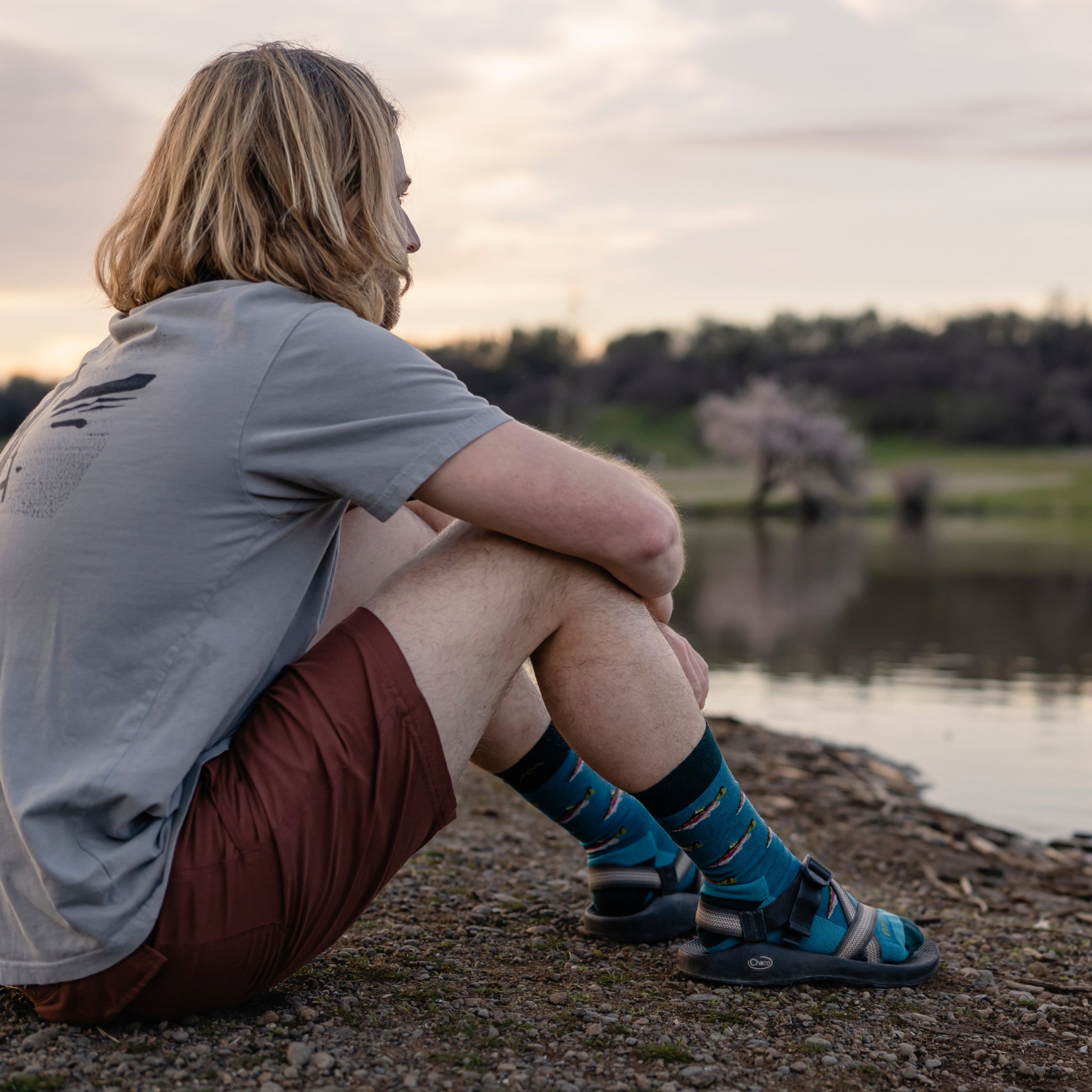 Model sitting by the edge of a lake wearing hiking sandals and 6085 socks in cascade colorway 