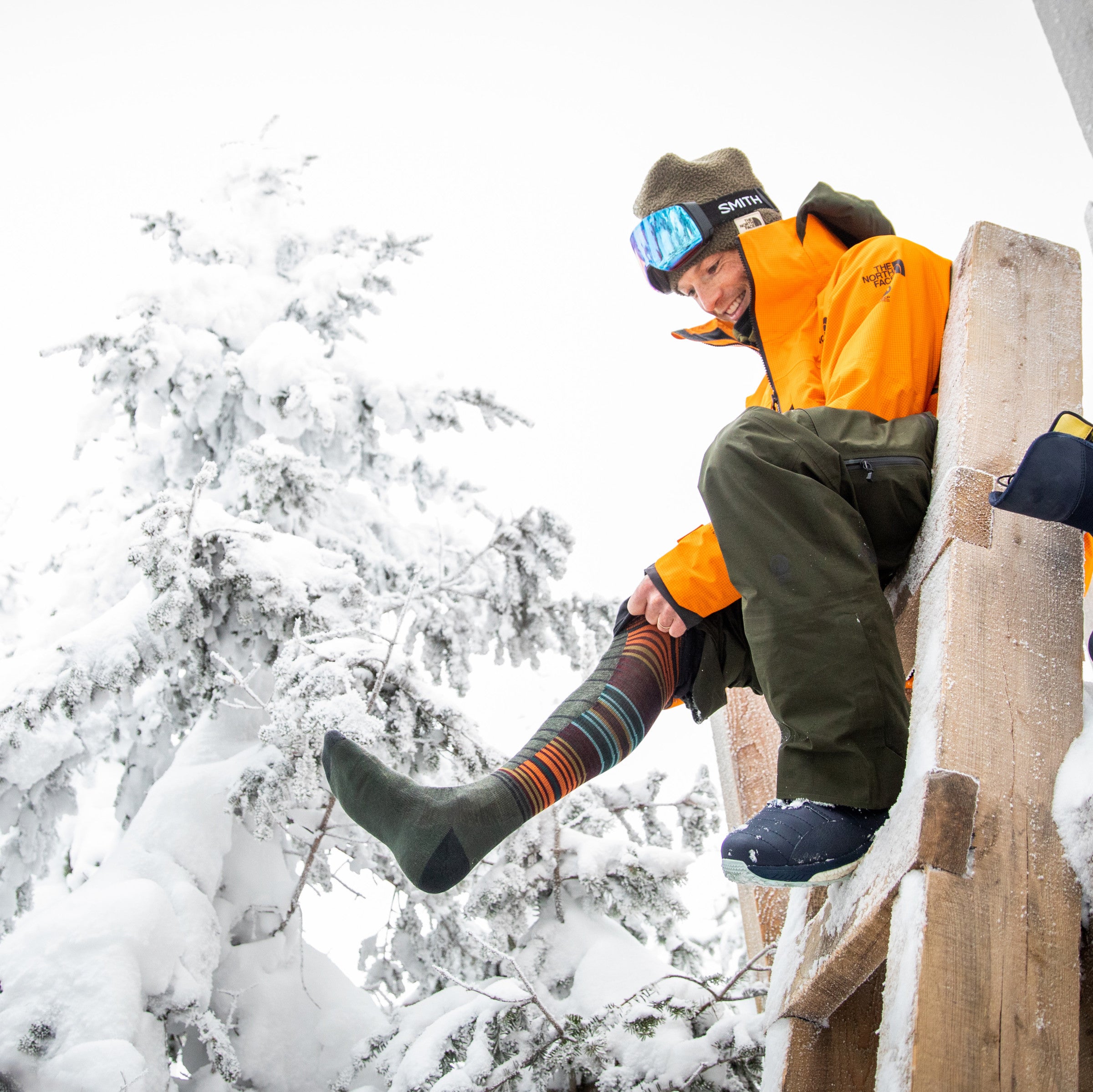 Full body shot of model sitting on a ladder outside in the snow wearing the men's backwoods over-the-calf snow sock in forest