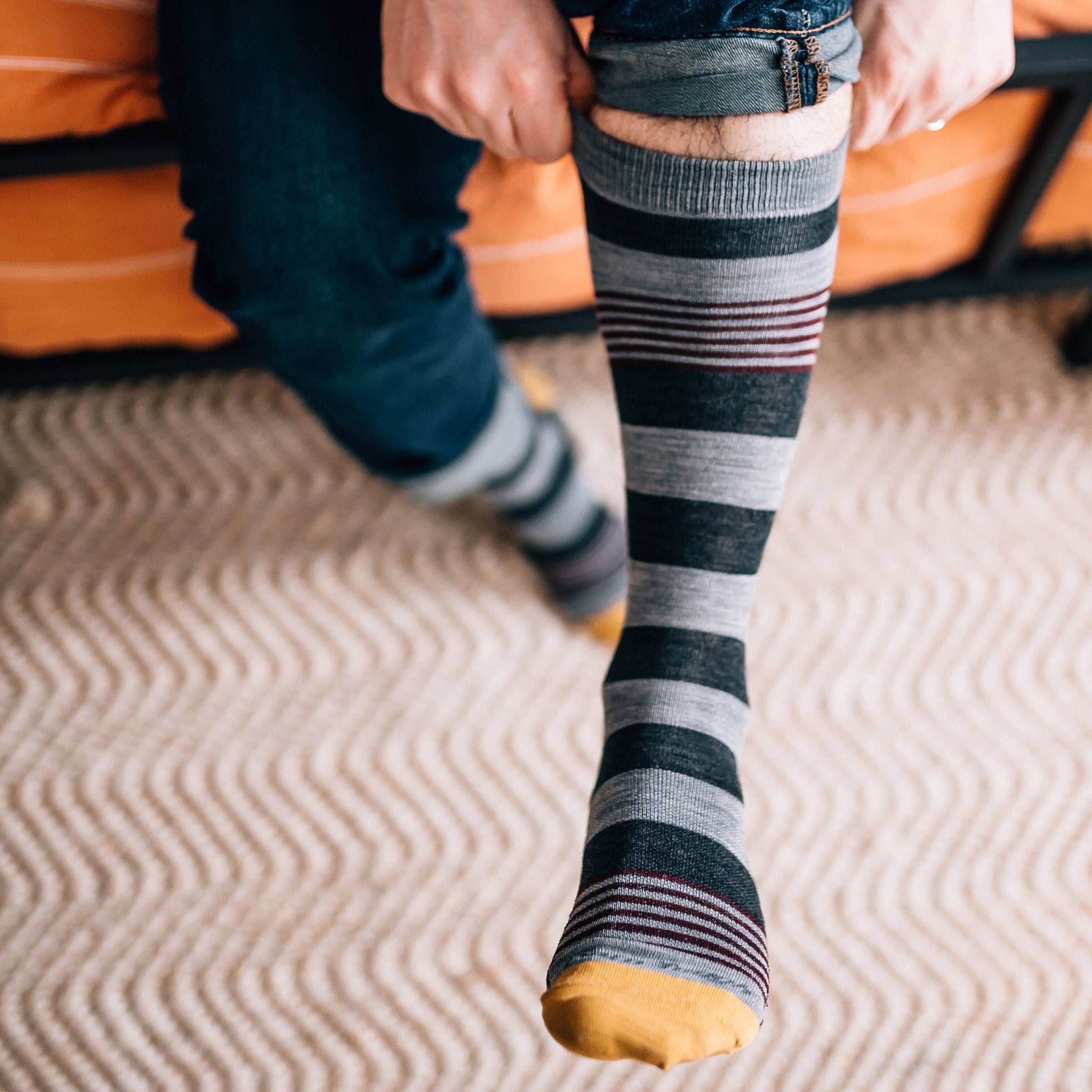 Man sitting on a couch, jeans rolled up pulling on his Oxford Crew Lightweight Lifestyle Socks in Gray