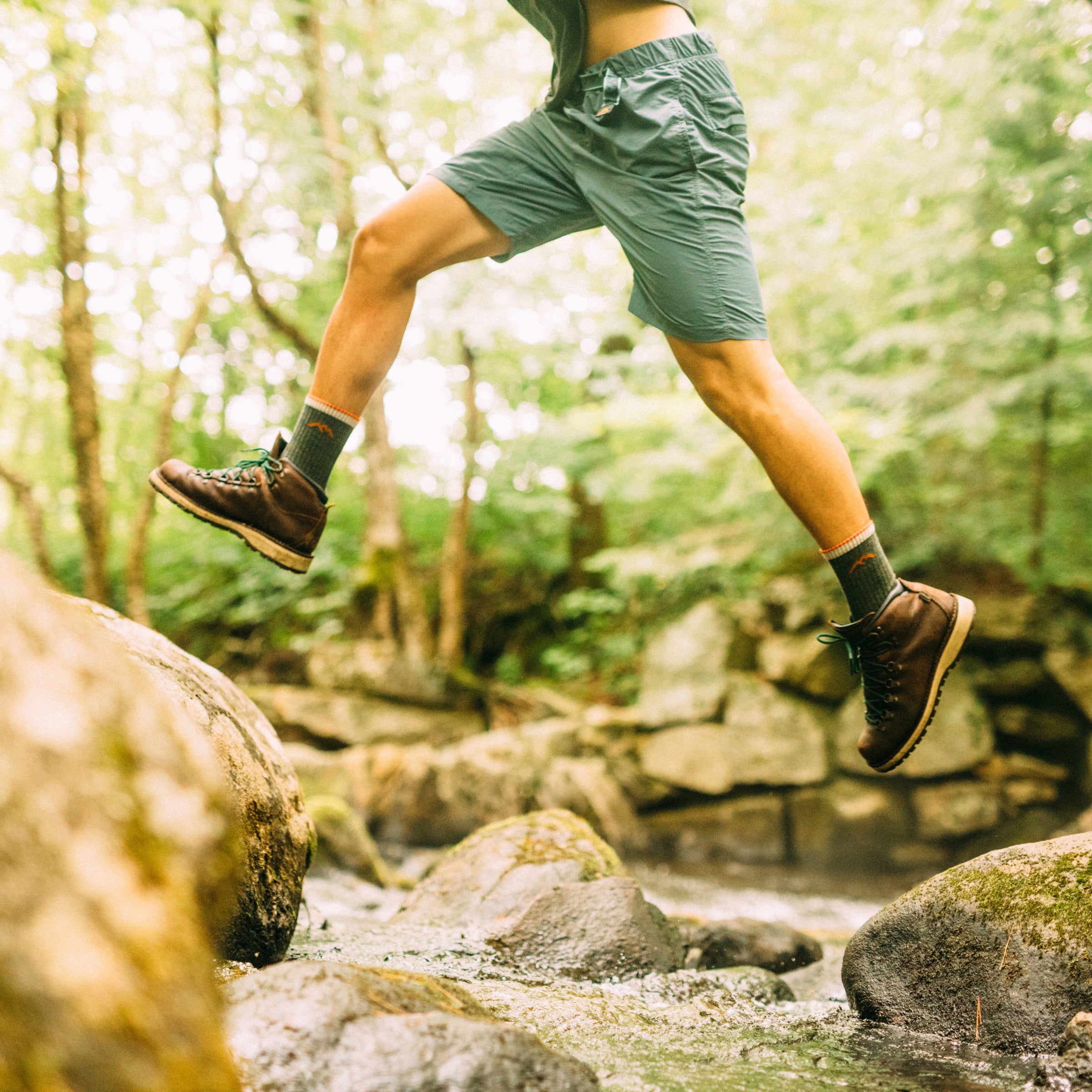 Side shot of model jumping over a river rock wearing the men's micro crew hiking sock in olive with brown shoes