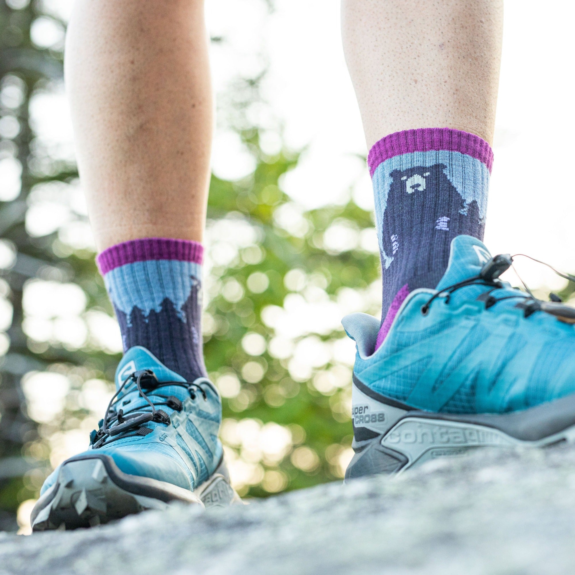 Close up shot of a model standing on a rock wearing the women's bear town micro crew hiking sock in purple with blue hiking shoes on