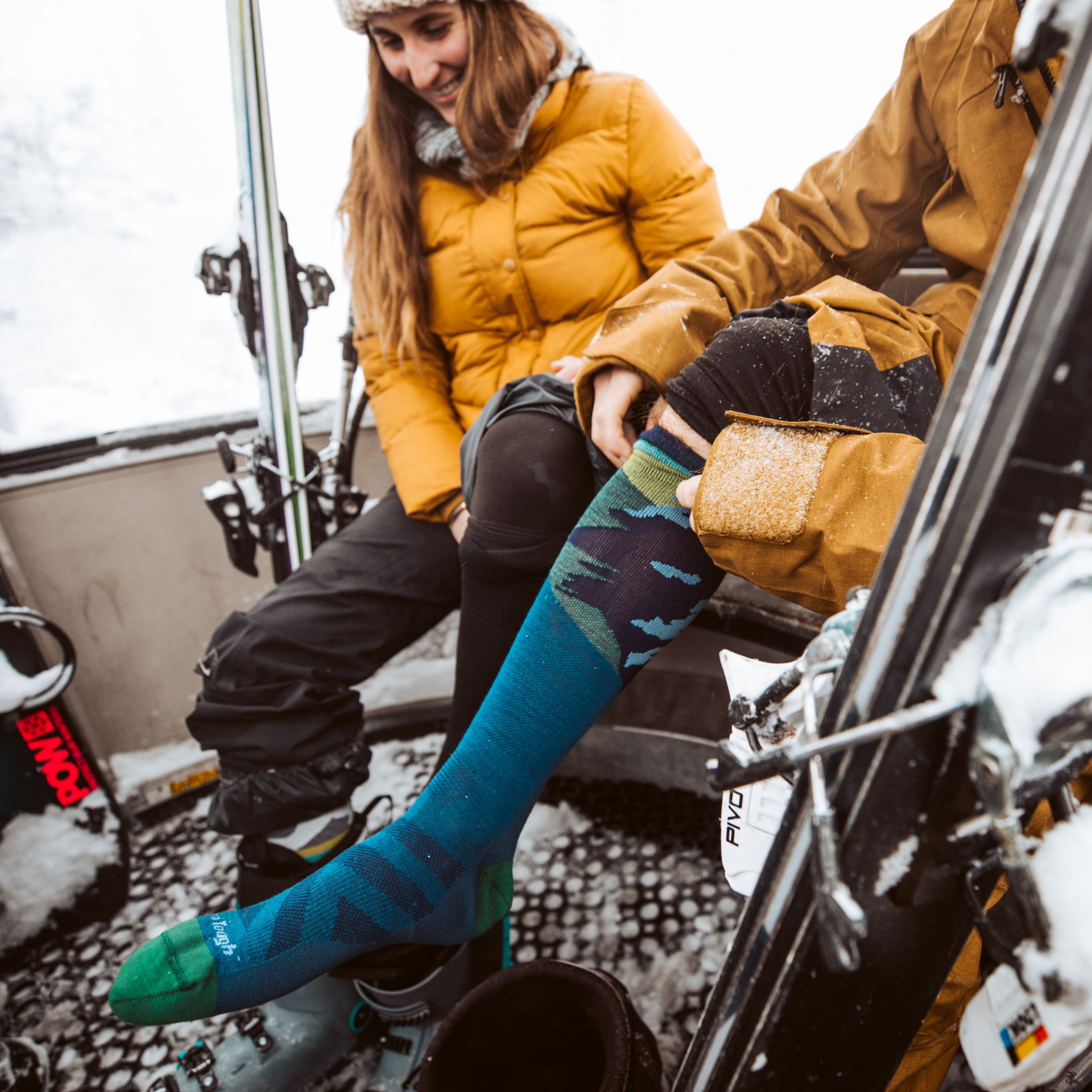 Side shot of model on a ski gondola wearing the men's solstice over-the-calf snow socks in midnight blue and a brown jacket