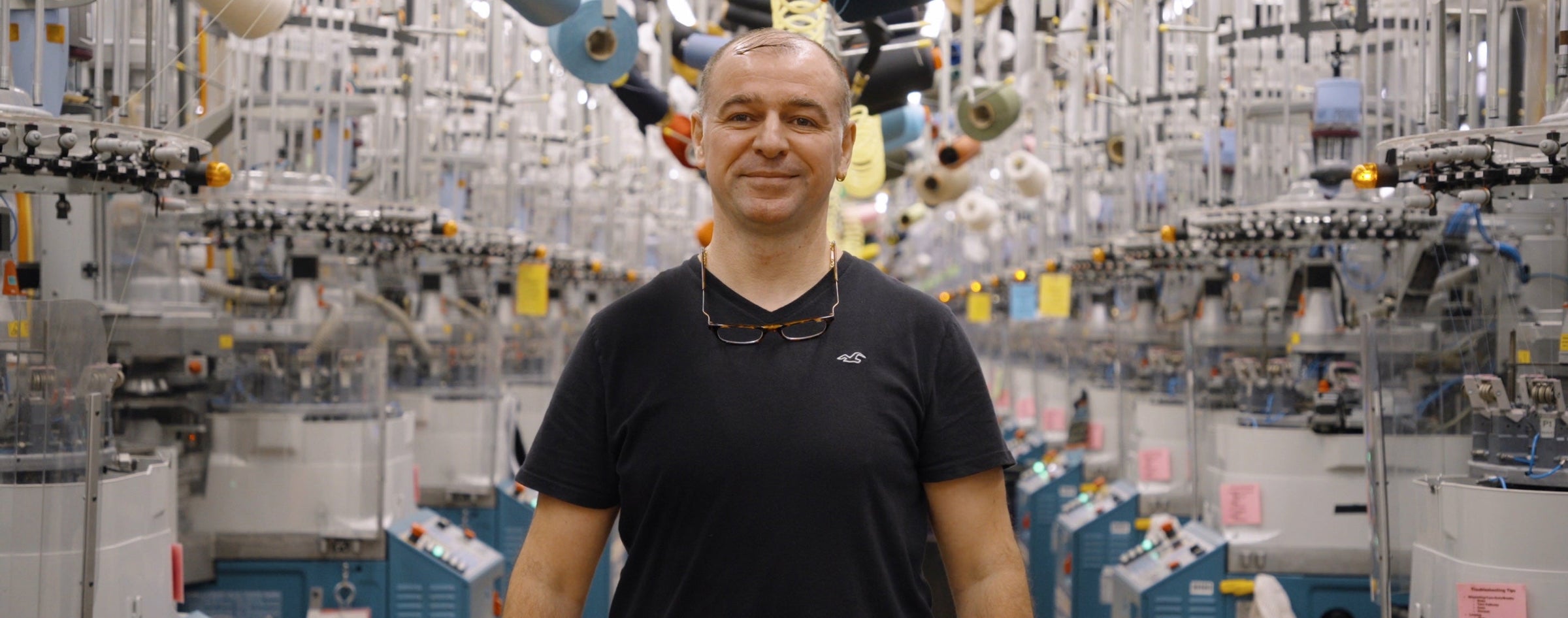 Wide cropped image of man standing on Darn Tough knitting floor with sock knitting machines on either side of him.