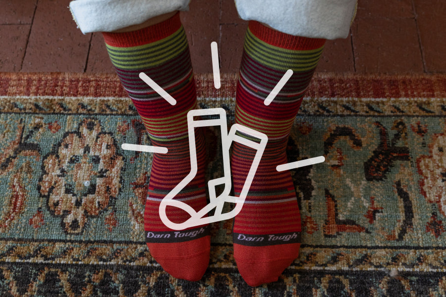 Feet on wearing red Darn Tough socks on green patterned rug with comfort sock graphic overlaid on image